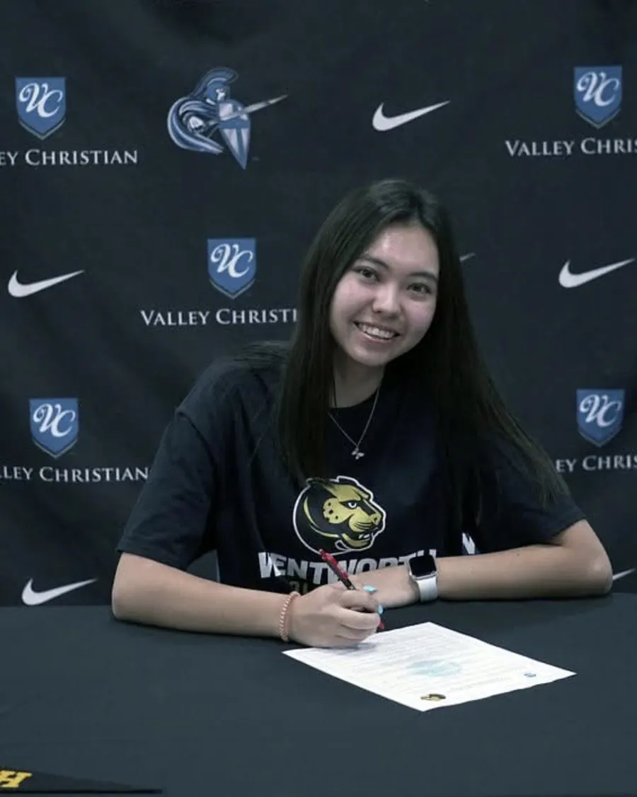 A student-athlete smiling as she signs her college letter at a Valley Christian signing-day table, wearing a Wentworth shirt.